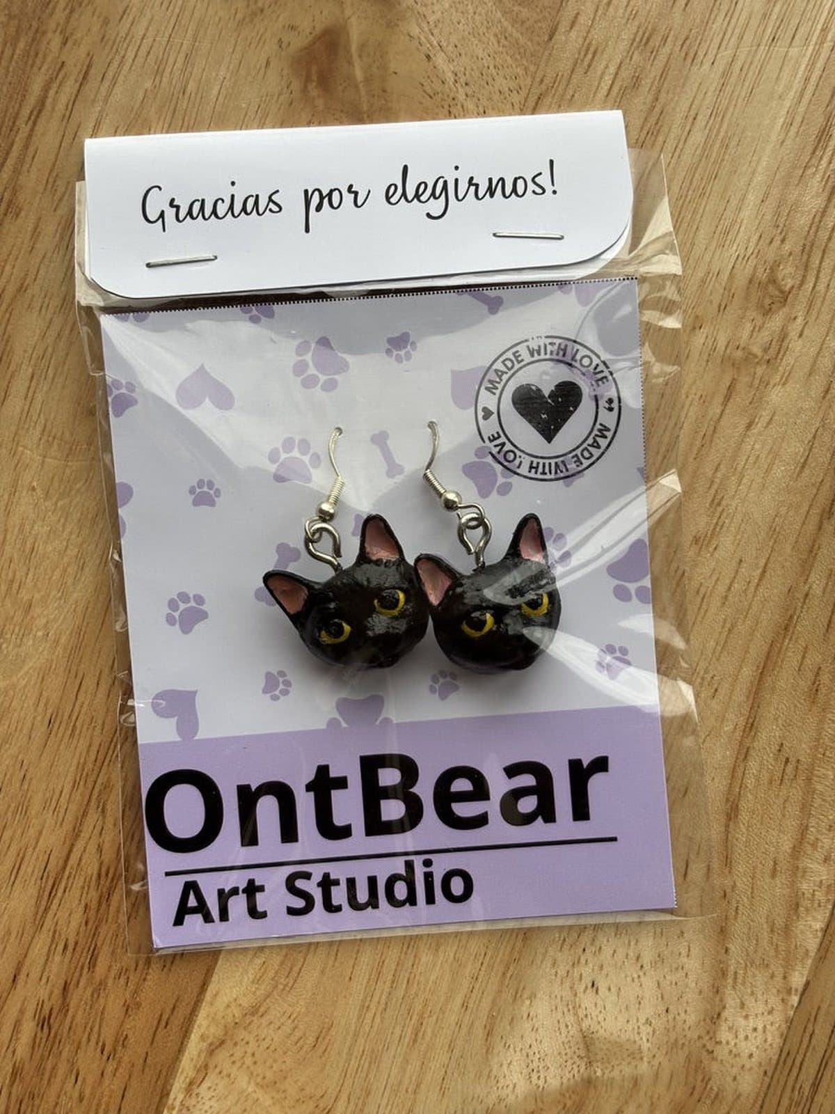 Par de aretes pintados a mano con caritas de gato negro de ojos amarillos, en su empaque de OntBear.