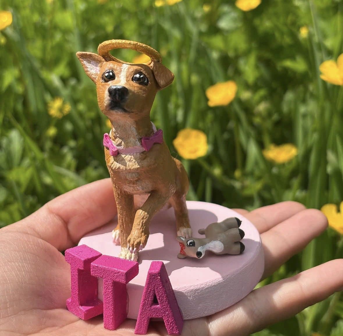 Figurita de un perro café con halo y moño rosa, sobre una base rosada con las letras ITA, sostenida en una mano entre flores amarillas.