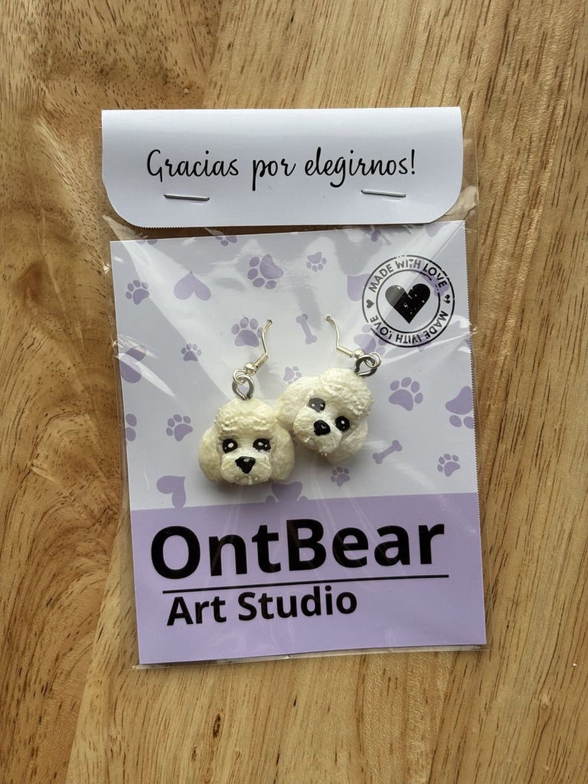 Par de aretes pintados a mano con caritas de caniche blanco, en su empaque de OntBear.