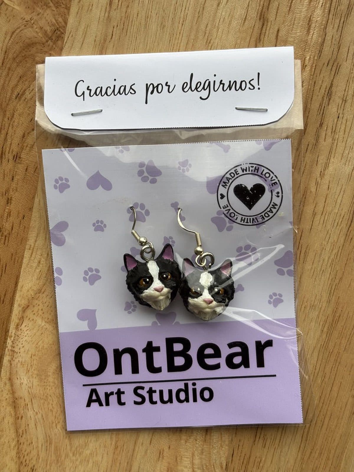 Par de aretes pintados a mano con caritas de gato negro y una franja blanca, en su empaque de OntBear.