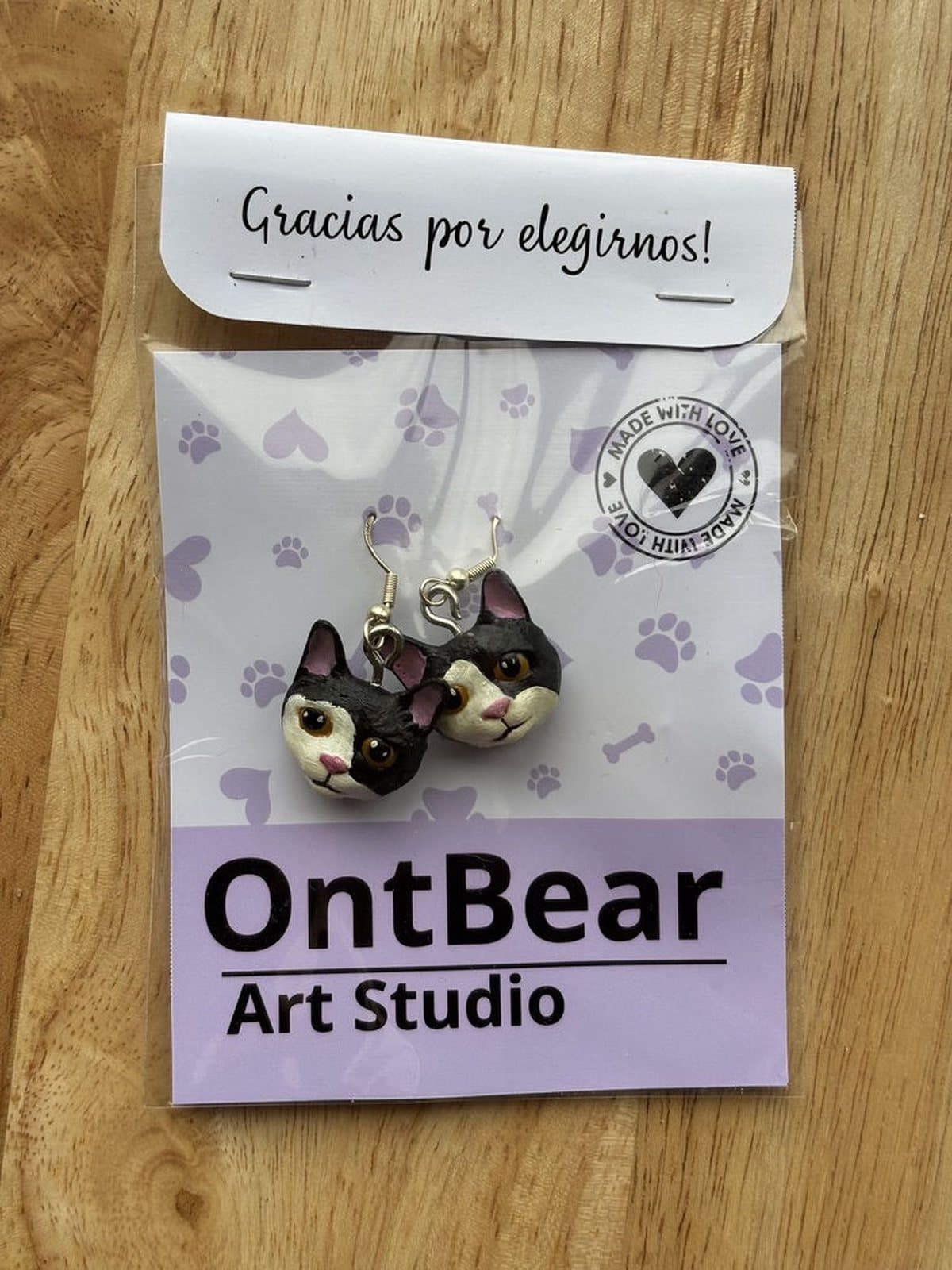 Par de aretes pintados a mano con caritas de gato blanco y negro, en su empaque de OntBear.