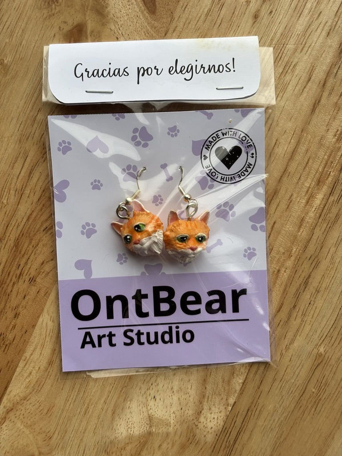 Par de aretes pintados a mano con caritas de gato naranjo y blanco, en su empaque de OntBear.