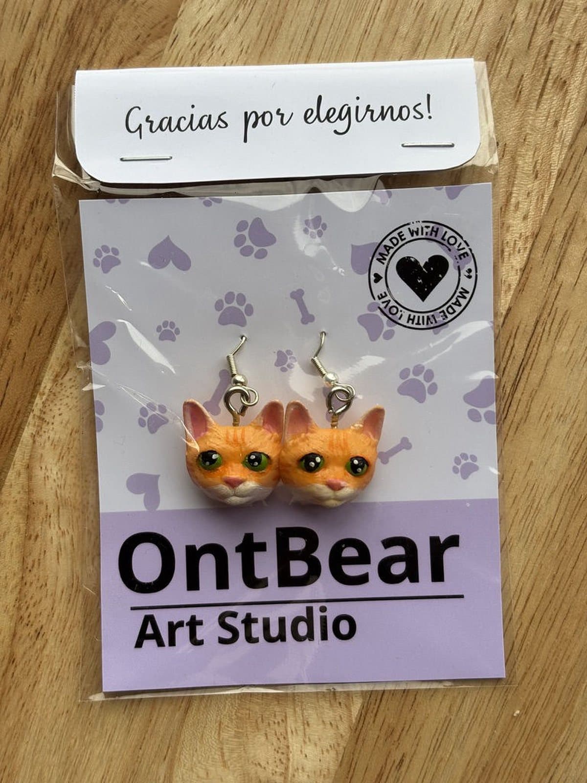 Par de aretes pintados a mano con caritas de gato naranjo de ojos verdes, en su empaque de OntBear.