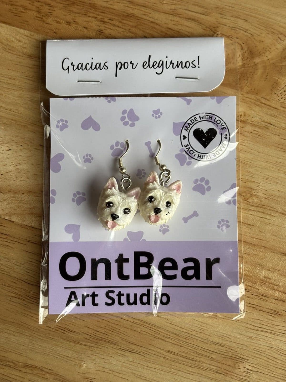 Par de aretes pintados a mano con caritas de westie y la lengua afuera, en su empaque de OntBear.