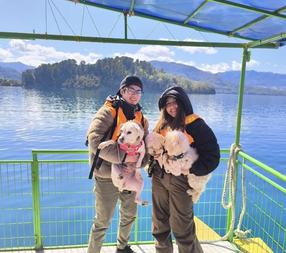 Catalina y Miguel sonriendo a bordo de una embarcación en un lago del sur, cada uno con sus perritos en brazos.