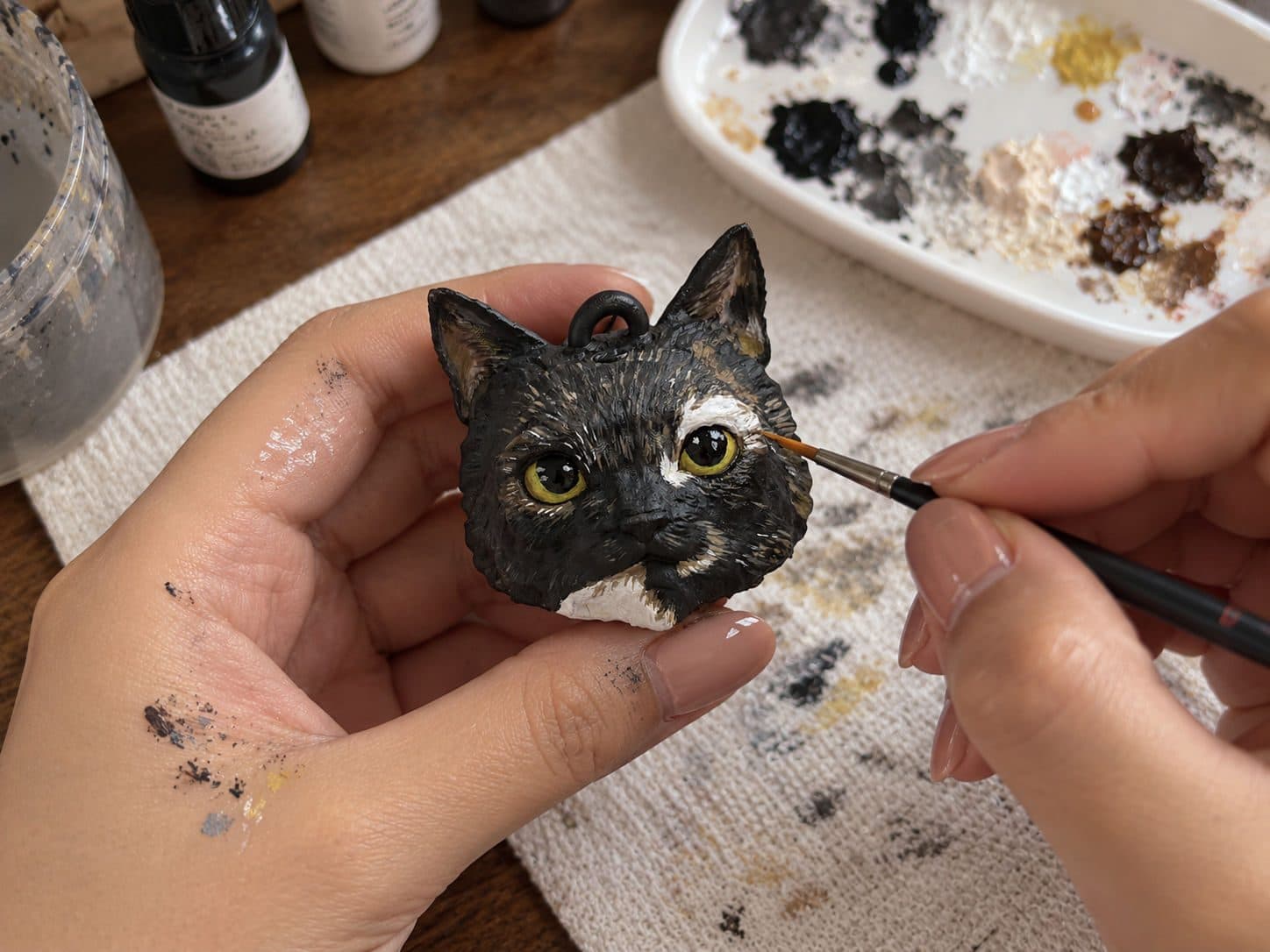 Las manos de Catalina pintan con un pincel fino los detalles del pelaje de la carita de una gata carey, junto a una paleta de pinturas.